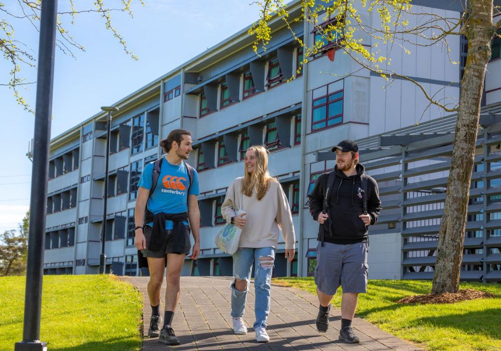 Three students walking along a path in front of a large building.